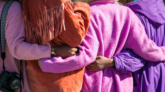 Fyra personer i lila kläder står bredvid varandra på rad och håller armarna runt varandras ryggar. Endast ryggen och armarna syns på personerna.